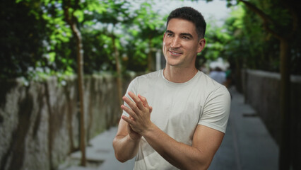 Fototapeta premium Young hispanic man clapping hands on a tree lined street with stone wall and dappled sunlight; encouragement optimism support.