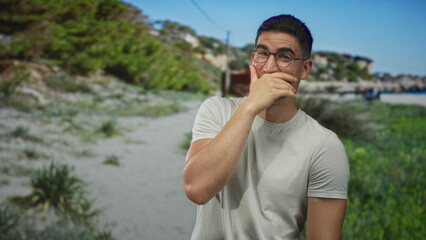 Man with hand covering mouth on street near seaside wearing glasses and tshirt, casual pose and...