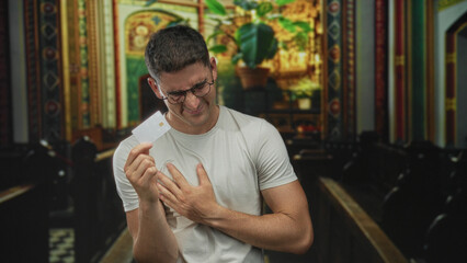 Young hispanic man holding a white card clutching chest for heartattack in a catholic church aisle; faith vulnerability.