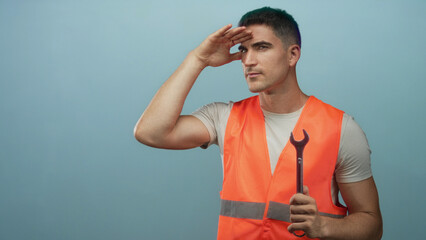 Man young hispanic in orange safety vest holding wrench and shading eyes looking into distance in blue studio; focus repair skill.