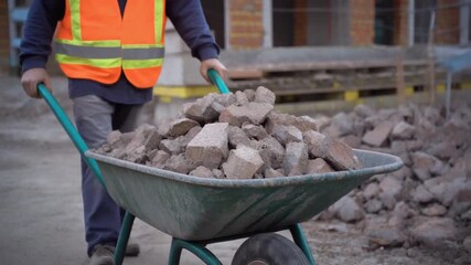 Construction worker carrying a wheelbarrow loaded with construction debris at construction site. The person is wearing safety vest