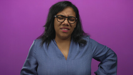 African american woman wearing glasses frowns and holds lower back in purple studio with eyes shut; backache.