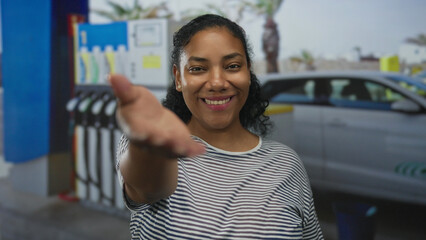 Smiling woman in striped shirt extends palm toward camera at petrol station forecourt by fuel pump and parked car  warm welcome. © Krakenimages.com