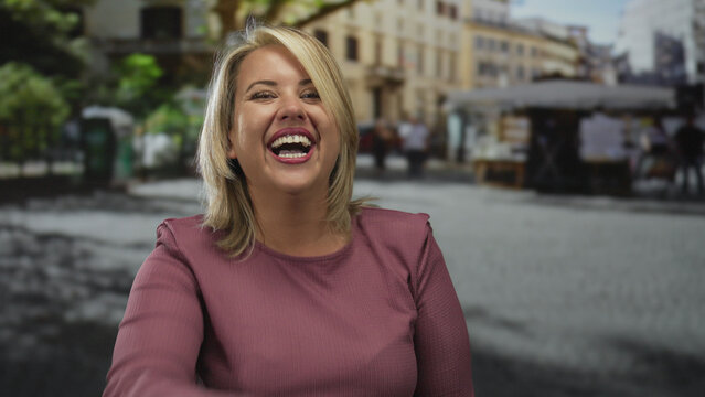 Woman laughs with hand on chest on a bustling city street with blurred buildings and moving pedestrians under bright daylight; joy.