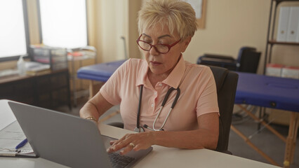 Woman physiotherapist typing on laptop with stethoscope around her neck and reading patient notes at desk in a medical building; calm professional care.