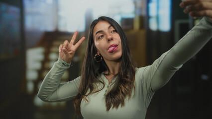 Young hispanic woman flashing peace sign and sticking out tongue with smartphone in hotel building;...