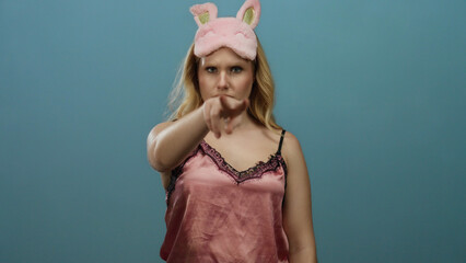 Young blonde woman in pink nightwear and bunny sleep mask gesturing while standing against a blue background, conveying a playful and watchful expression.