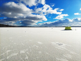 Ice fishing tent stands on a frozen lake under a blue sky with clouds in winter