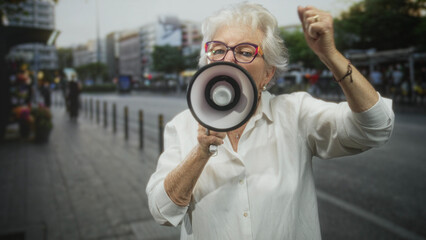 Senior woman wearing white blouse and glasses shouts into megaphone and raises fist on a busy city...