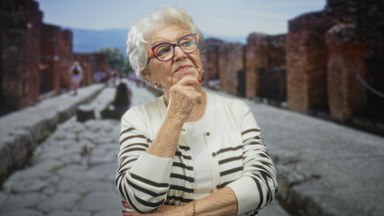 Senior woman with hand on chin wearing colorful glasses and striped cardigan, posed on a cobbled...