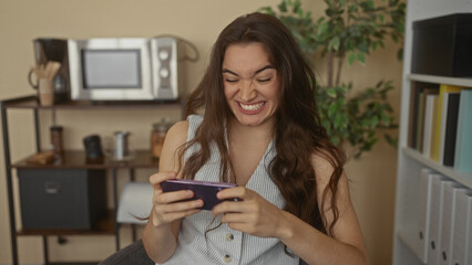 Young hispanic woman holds smartphone with both hands and pumps fist high in office space; excitement.