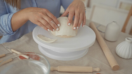 Woman hands smoothing white clay over a rotating wheel platform in a sunlit studio workspace;...