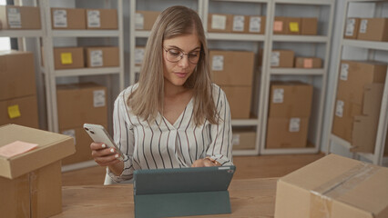 Woman sits at desk surrounded by packages while holding smartphone and tapping tablet in building;...