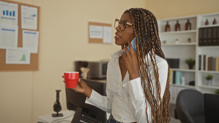Woman with long braids and glasses holding phone to ear and a red mug near desk and monitor in building; focused workday.