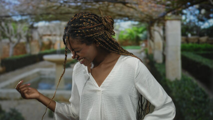 Woman laughing with hands raised near a stone fountain under a pergola in a forest garden with...