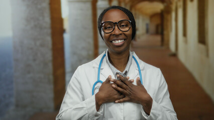 Woman doctor standing confidently in a historical university hallway with stethoscope around neck while smiling, exhibiting pride and assurance in her academic environment.