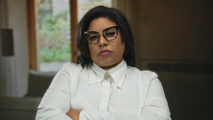 Woman indoors at hotel, with crossed arms, wearing glasses, staring confidently, background showing...