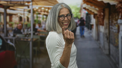 Woman grey haired middle aged wearing glasses beckoning with hand and forearm visible at outdoor restaurant terrace on a busy street; playful invitation.