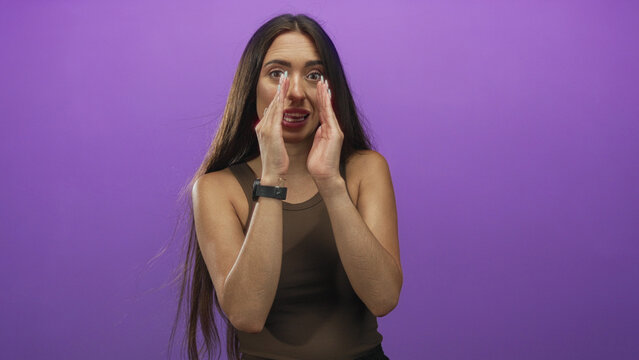 Young hispanic woman cupping hands to mouth in purple studio wearing brown tank top and smartwatch; urgent message.