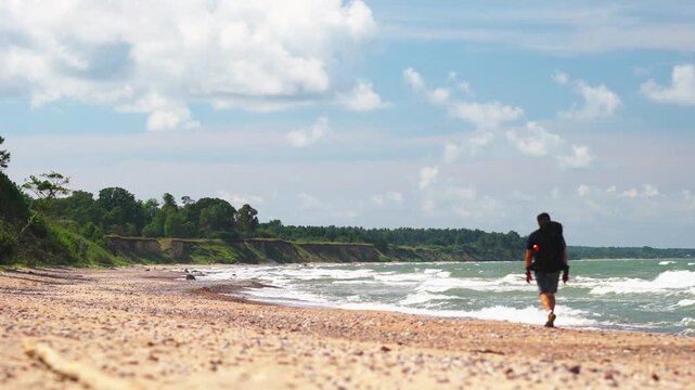 man walking on sandy beach under blue sky, small backpack, footprints along wet sand, gentle waves breaking on shore. Camera moving to the left
