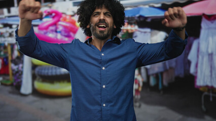 Man with curly hair raising fists on street market aisle beside souvenir stalls, smiling and shouting in open air; joy celebration.
