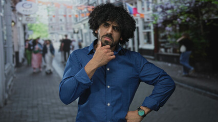 Man with hand on chin on street, curly hair and beard, gesturing with free hand while pausing to...