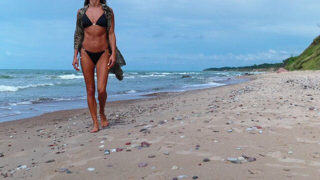 woman in black bikini walking toward camera on sandy beach, confident stride with sunglasses, closeup details of legs.