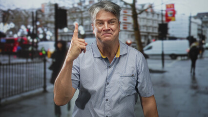 Middle aged man pointing index finger upward on busy wet street, smiling in short sleeve shirt and...