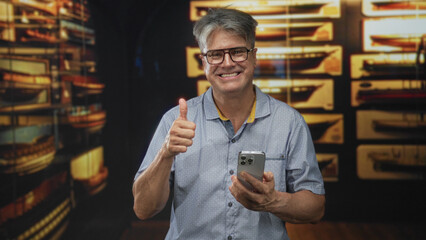 Man holding smartphone tapping screen and giving thumbs up in building, smiling while focused on...