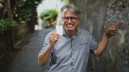 Senior man holding creditcard and pointing finger in narrow street, smiling and sticking tongue out while presenting the chip card; playful confidence.