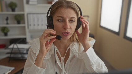 Woman adjusting headset and microphone at office desk with hands near ears and mouth, looking at computer monitor while speaking and listening through headset; concentration productivity.