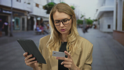 Fototapeta premium Woman using tablet and card on city street, focused expression in outdoor urban setting.