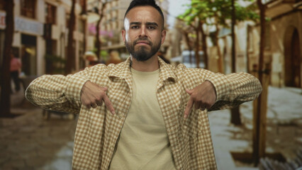 Man pointing both index fingers down toward his torso on a street with checkered shirt and beard; self assertion defiance.