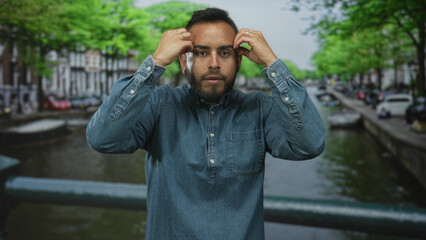 Man touching his face with both hands on a canal bridge on a street in amsterdam, eyes closed and steady expression; quiet contemplation.