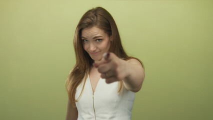 Woman points finger to temple in a green studio while wearing a white sleeveless top and tilting head; uncertainty doubt introspection.