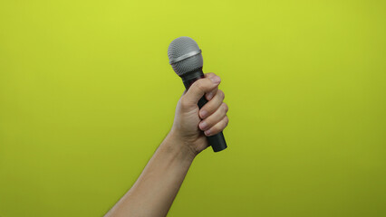 Man holding microphone against vibrant yellow wall, showcasing male hand gripping mic with green...