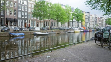 Obraz premium Blurred view of amsterdam's iconic canals with classic buildings and moored boats casting reflections on water on a calm day.