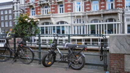 Obraz premium Bicycles parked beside a canal in amsterdam with blurred historic architecture and vibrant flowers in the background, creating a serene urban scene.