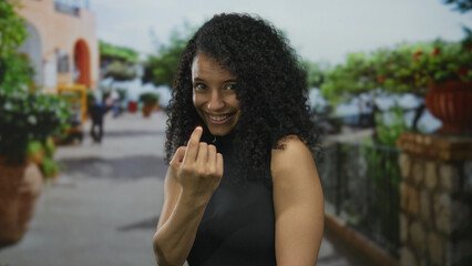 Fototapeta premium Smiling young hispanic woman beckons with finger on a sunlit outdoor street; playfulness invitation.
