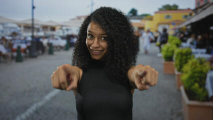 Hispanic woman with curly hair pointing finger to camera and smiling on sunlit street; playful invitation.