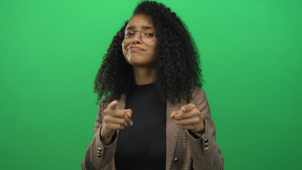 Woman smiling in glasses and plaid jacket, pointing fingers confidently at the camera with a playful expression on an isolated green background.