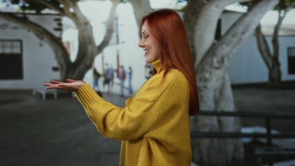 Woman wearing yellow sweater extends hands on city street with blurred background showcasing trees and architecture.