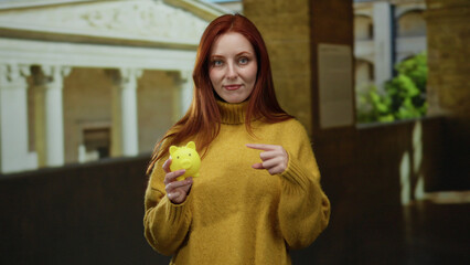 Woman holding yellow piggy bank in front of ancient building, wearing warm sweater with red hair, embodying finance and education themes outdoors © Krakenimages.com