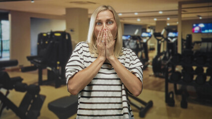 Woman covers mouth with both hands in gym among dumbbells and treadmills, striped shirt and visible...