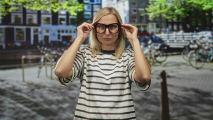 Woman adjusting thick glasses with both hands on frame on street by bicycles and canal, striped shirt visible; contemplation.