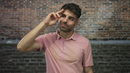 Young hispanic man in pink shirt points finger to temple in studio against brick wall; thoughtful reflection.
