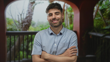 Young hispanic man with arms crossed in building near a lush pool deck under tropical foliage;...