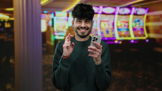 Young man crossing fingers holds smartphone in a lively casino setting with colorful slot machines in the background, conveying hope and excitement. - Powered by Adobe
