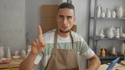 Man in apron points index finger upward while seated at a workbench with pottery shelves in a studio; craft concentration.