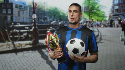 Man holding gold trophy and soccer ball with both hands on cobbled canal street, wearing blue and...